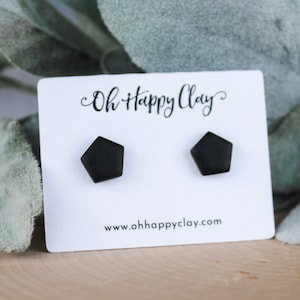 May include: A pair of black, pentagon-shaped stud earrings displayed on a white card. The card features the text "Oh Happy Clay" in a cursive font and the website address. The earrings are set against a backdrop of green foliage.