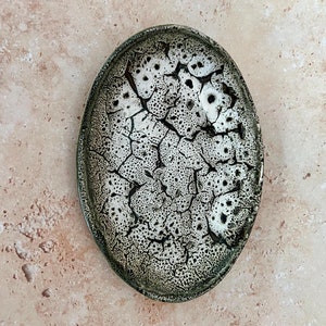 Pode incluir: Um prato oval de cerâmica branca com um esmalte verde e um acabamento rachado. O esmalte tem um padrão de pequenos pontos e linhas pretas.