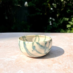 Op de afbeelding: Een kleine, witte keramische kom met een abstract ontwerp in groen en wit. De kom staat op een lichtbruin oppervlak.