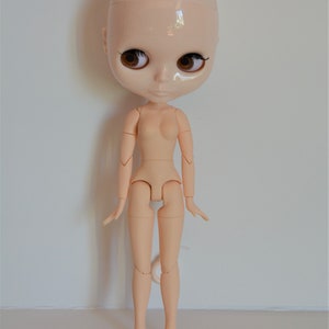 May include: A nude-coloured articulated doll with brown eyes and no hair. The doll is standing with its arms by its sides and its legs slightly apart.