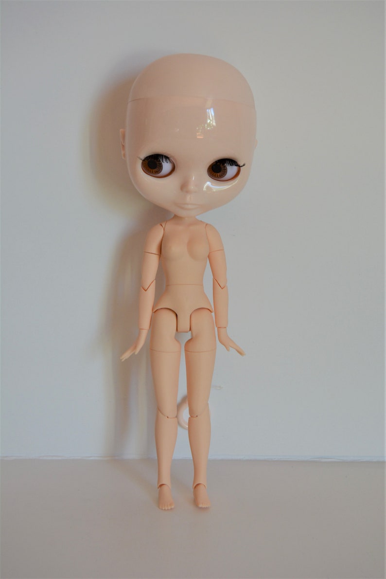 May include: A nude-coloured articulated doll with brown eyes and no hair. The doll is standing with its arms by its sides and its legs slightly apart.