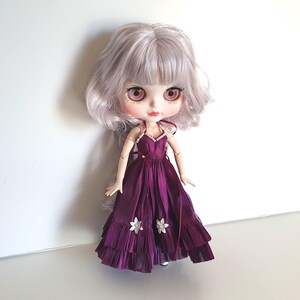 K&ouml;nnte beinhalten: Eine Puppe mit langen, welligen, silbernen Haaren, die ein violettes Kleid mit wei&szlig;en Blumen tr&auml;gt. Die Puppe hat gro&szlig;e, rosafarbene Augen und einen rosa Rouge auf den Wangen.