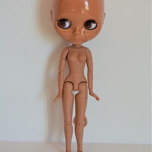 May include: A nude-coloured articulated doll with brown eyes and a bald head. The doll is standing with its arms by its sides and its legs slightly apart.