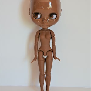 May include: A brown articulated doll with a bald head and brown eyes. The doll is posed standing with its arms outstretched.