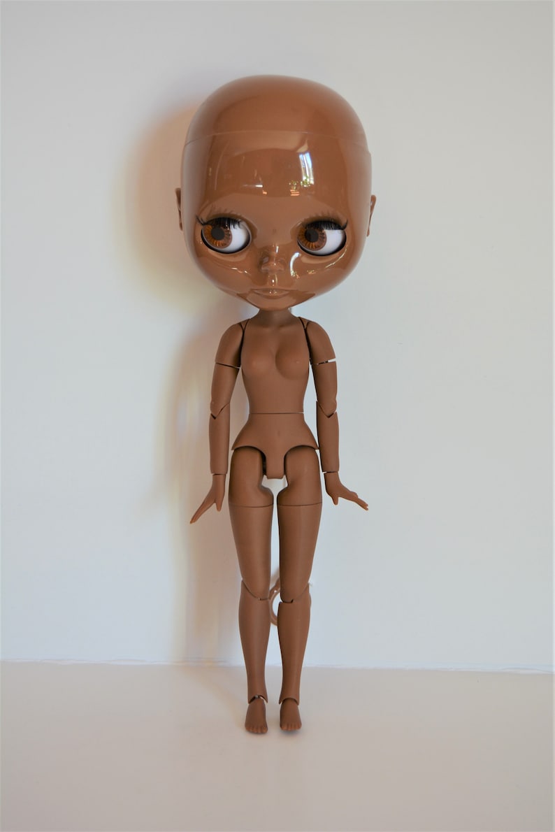 May include: A brown articulated doll with a bald head and brown eyes. The doll is posed standing with its arms outstretched.