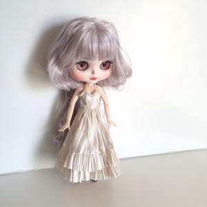 K&ouml;nnte beinhalten: Eine Puppe mit langen, welligen, silbernen Haaren, die ein wei&szlig;es Kleid mit einem R&uuml;schen-Saum tr&auml;gt.