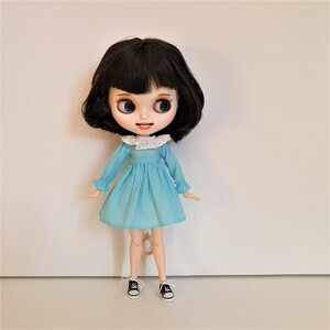 Puede incluir: Una mu&ntilde;eca con cabello negro y ojos azules que lleva un vestido azul claro con un cuello blanco y zapatos negros.