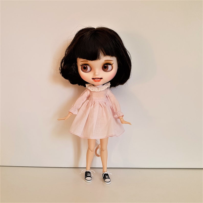 Puede incluir: Una mu&ntilde;eca con cabello negro y ojos marrones que lleva un vestido rosa con un cuello de encaje blanco. La mu&ntilde;eca lleva zapatillas negras y blancas.