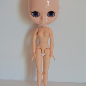 May include: A nude-coloured articulated doll with blue eyes and no hair. The doll has a realistic face and is posed standing with its arms by its sides.