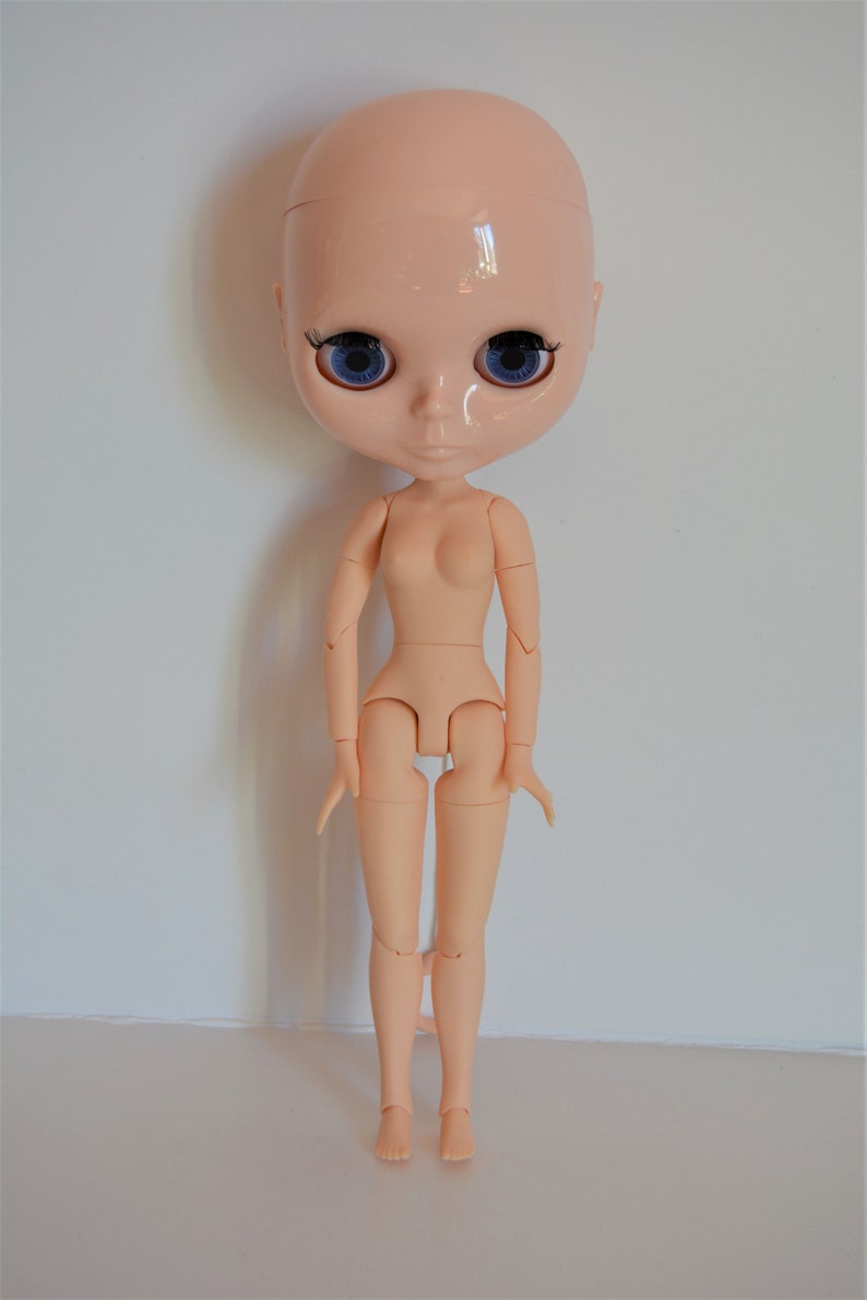 May include: A nude-coloured articulated doll with blue eyes and no hair. The doll has a realistic face and is posed standing with its arms by its sides.