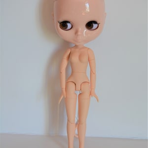May include: A nude-coloured articulated doll with brown eyes and a bald head. The doll is standing with its arms by its sides and its legs slightly apart.