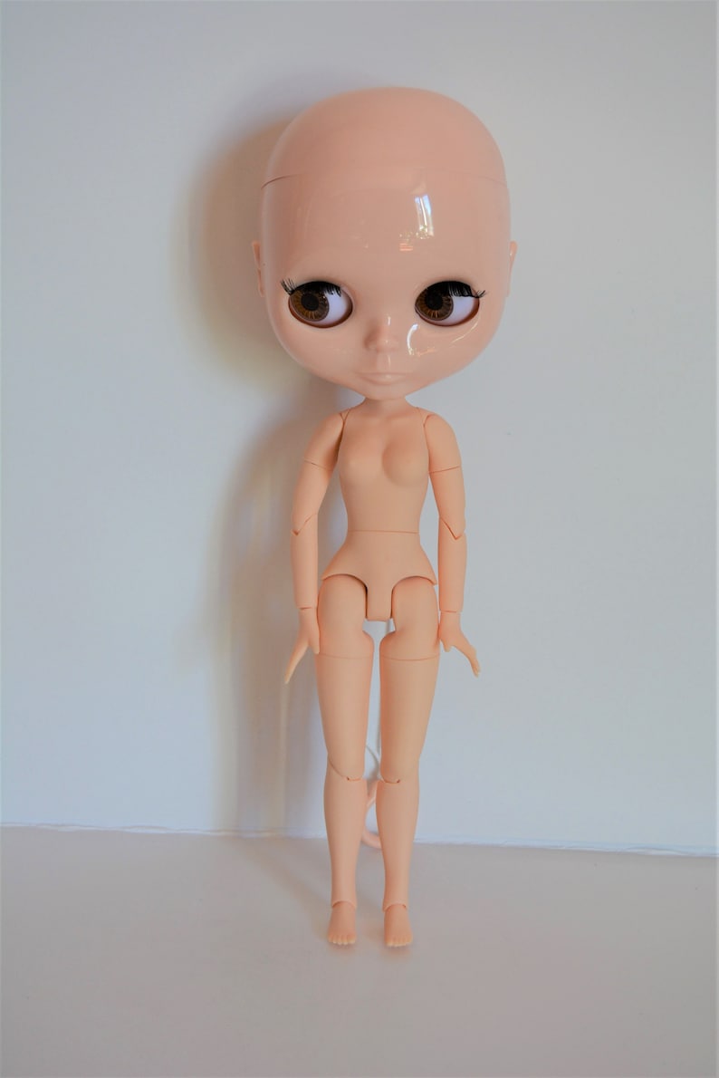 May include: A nude-coloured articulated doll with brown eyes and a bald head. The doll is standing with its arms by its sides and its legs slightly apart.