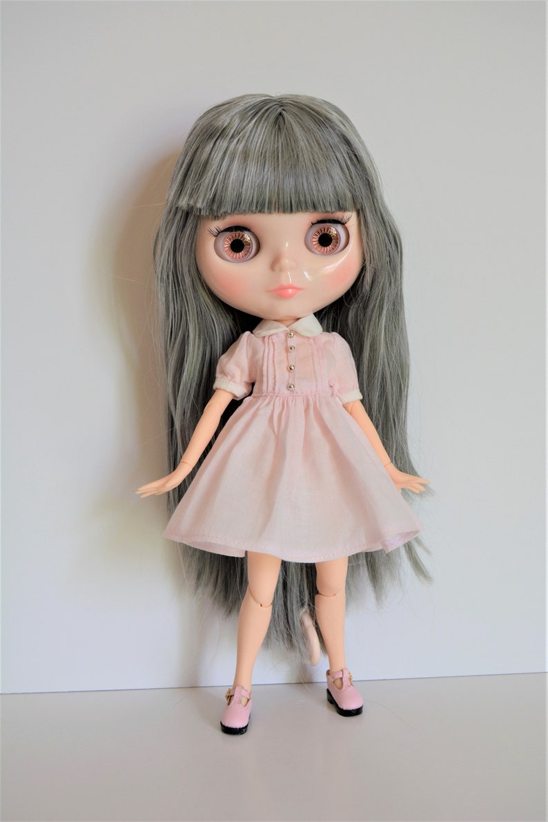 May include: A doll with long gray hair, wearing a pink dress with a white collar and pink shoes.