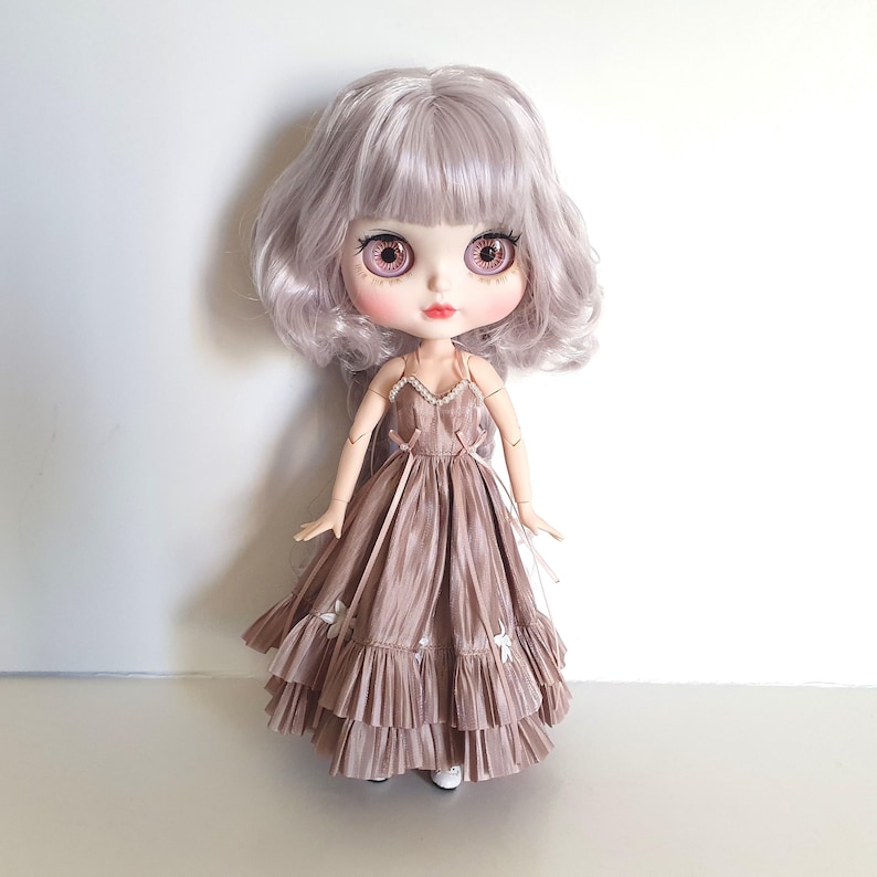 K&ouml;nnte beinhalten: Eine Puppe mit langen, welligen, silbernen Haaren, die ein braunes Kleid mit wei&szlig;en Akzenten tr&auml;gt. Das Kleid hat einen ger&uuml;schten Rock und einen herzf&ouml;rmigen Ausschnitt.