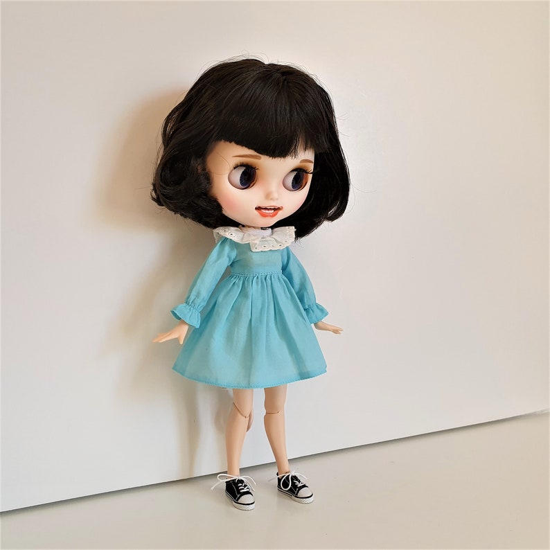 Puede incluir: Una mu&ntilde;eca con cabello negro y ojos azules que lleva un vestido azul claro con un cuello de encaje blanco y zapatos negros.