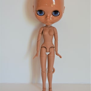 May include: A nude-coloured articulated doll with blue eyes. The doll has a bald head and is posed standing with its arms by its sides.