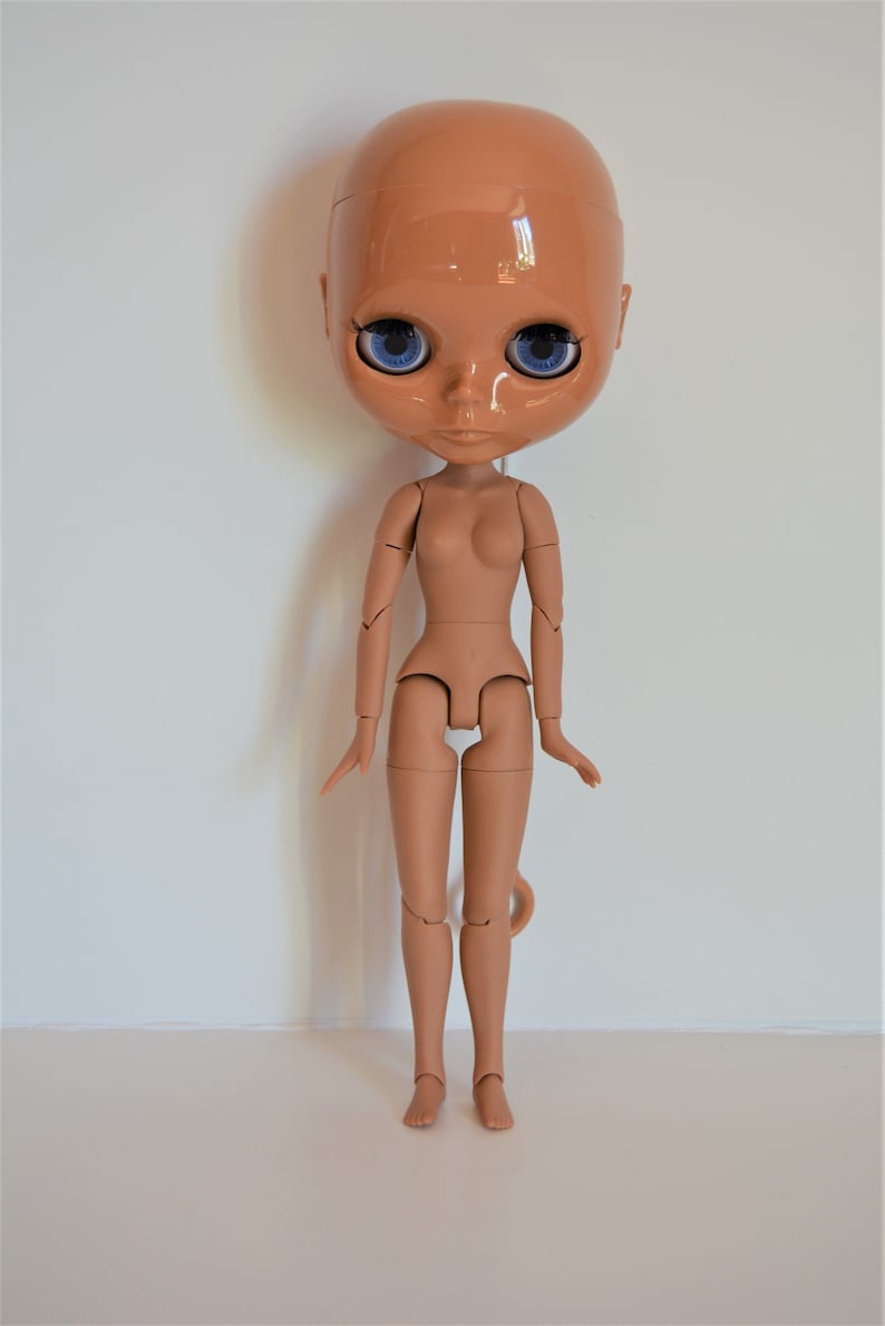 May include: A nude-coloured articulated doll with blue eyes. The doll has a bald head and is posed standing with its arms by its sides.