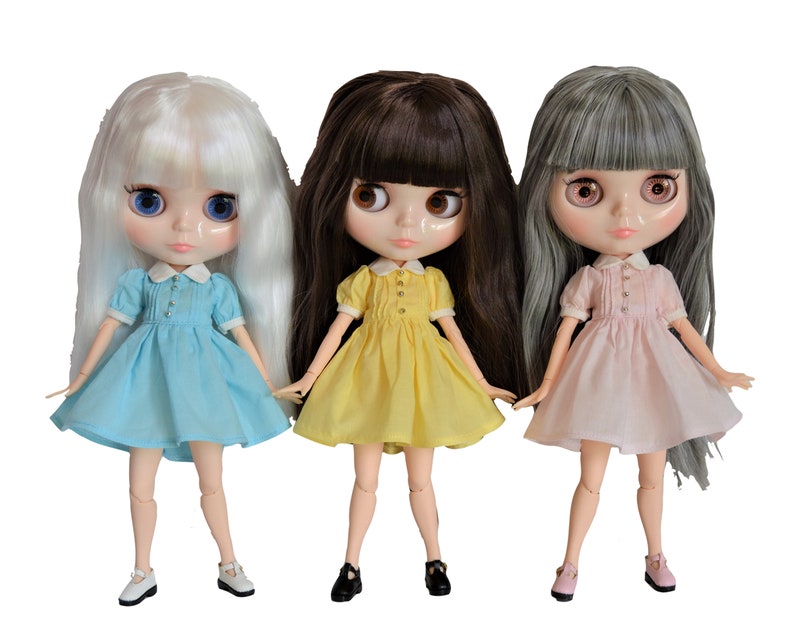 May include: Three dolls with long hair wearing different colored dresses. The doll on the left has white hair and is wearing a light blue dress. The doll in the middle has brown hair and is wearing a yellow dress. The doll on the right has gray hair and is wearing a pink dress.