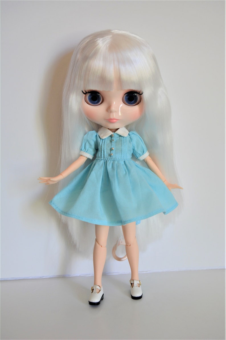 May include: A white doll with long, straight, white hair wearing a light blue dress with a white collar and white shoes.