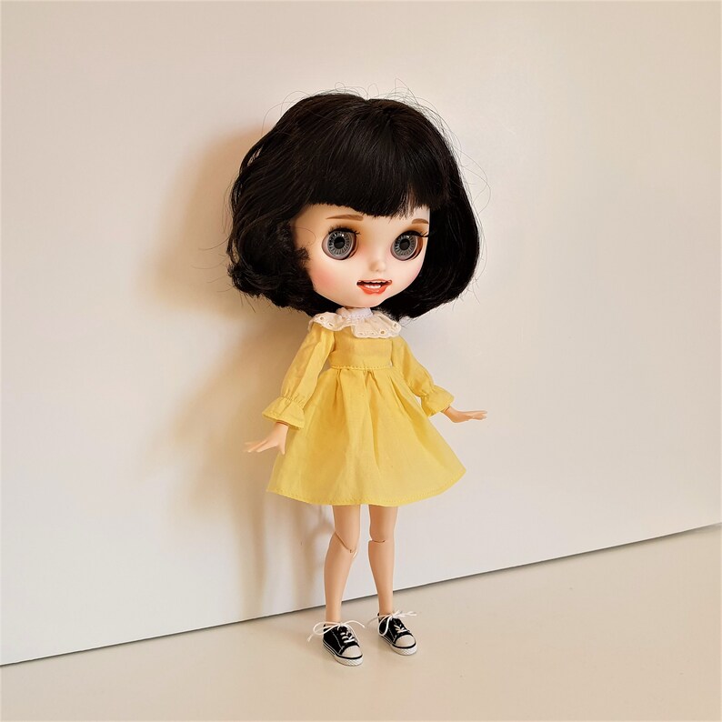 Puede incluir: Un vestido amarillo con un cuello de encaje blanco y zapatos negros. La mu&ntilde;eca tiene el pelo negro y los ojos azules.