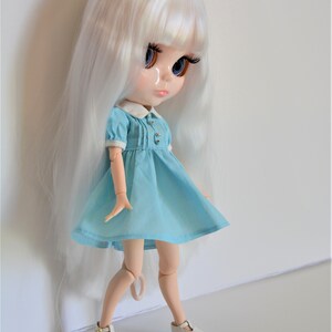 May include: A white haired doll wearing a light blue dress with white trim and white shoes. The doll has blue eyes and a painted smile.