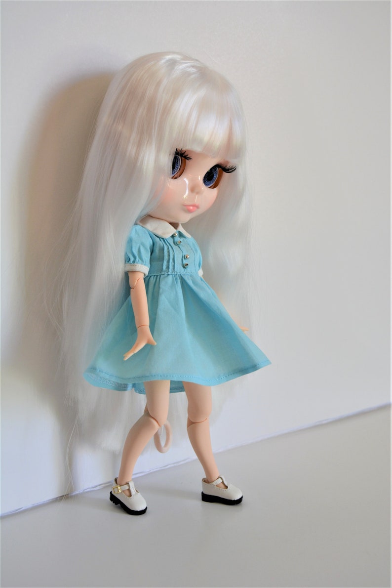 May include: A white haired doll wearing a light blue dress with white trim and white shoes. The doll has blue eyes and a painted smile.