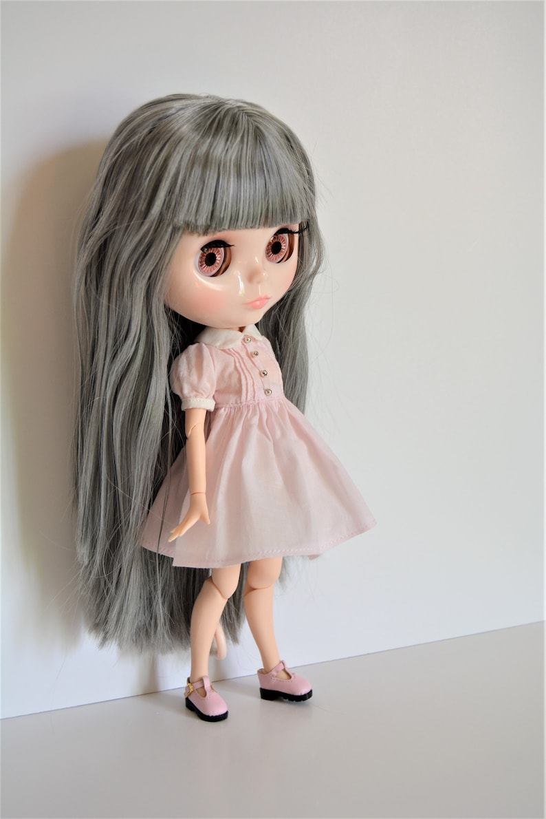 May include: A doll with long gray hair, wearing a pink dress with a white collar and pink shoes.
