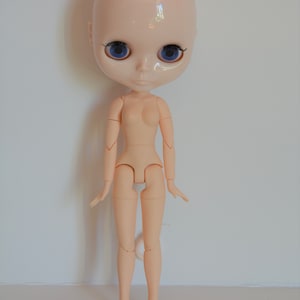 May include: A nude-coloured doll with blue eyes and a bald head. The doll has articulated limbs and is posed standing with its arms slightly raised.