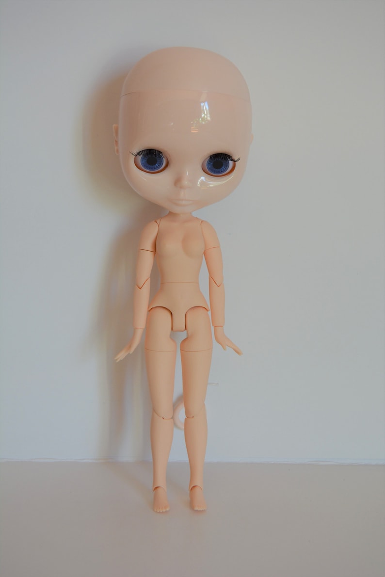 May include: A nude-coloured doll with blue eyes and a bald head. The doll has articulated limbs and is posed standing with its arms slightly raised.