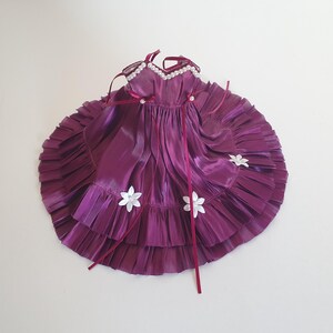 K&ouml;nnte beinhalten: Ein lilafarbenes Satin-Puppenkleid mit wei&szlig;en Blumenverzierungen und einem Perlenbesatz am Ausschnitt.