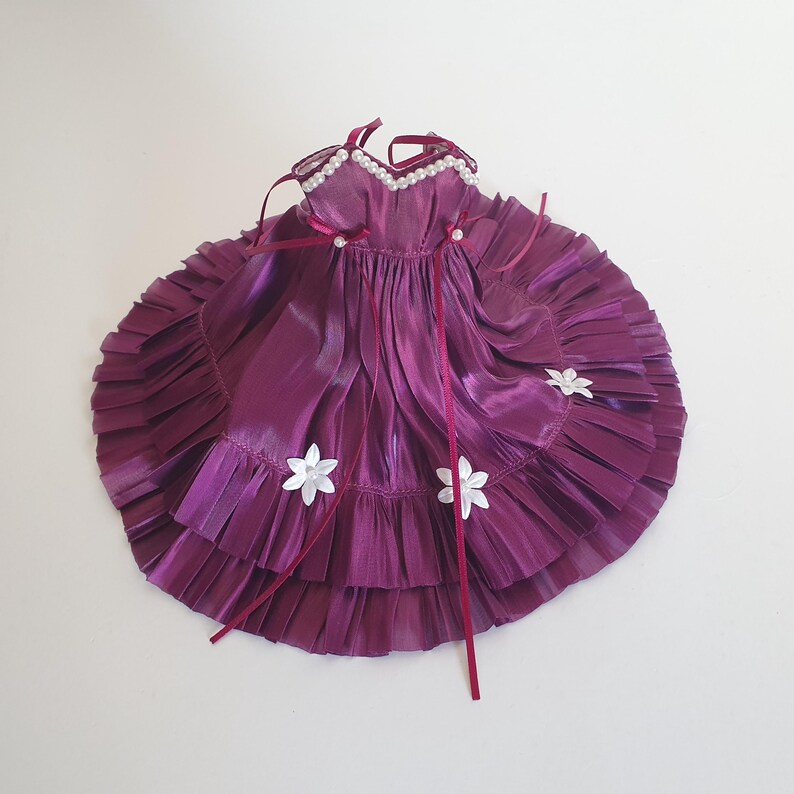 K&ouml;nnte beinhalten: Ein lilafarbenes Satin-Puppenkleid mit wei&szlig;en Blumenverzierungen und einem Perlenbesatz am Ausschnitt.
