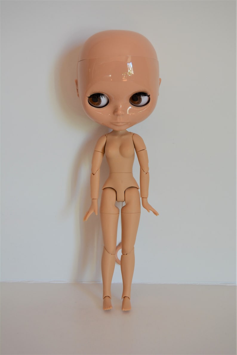 May include: A nude-coloured articulated doll with brown eyes and a bald head. The doll is posed standing with its arms at its sides and its legs slightly apart.