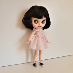 Puede incluir: Una mu&ntilde;eca con cabello negro y ojos marrones que lleva un vestido rosa con un cuello de encaje blanco y zapatos negros.