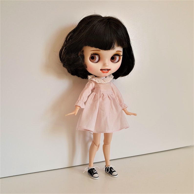 Puede incluir: Una mu&ntilde;eca con cabello negro y ojos marrones que lleva un vestido rosa con un cuello de encaje blanco y zapatos negros.