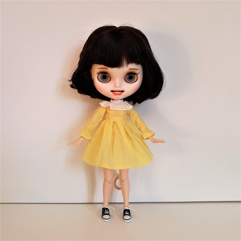 Puede incluir: Un vestido amarillo con un cuello de encaje blanco y zapatos negros. La mu&ntilde;eca tiene el pelo negro y los ojos azules.