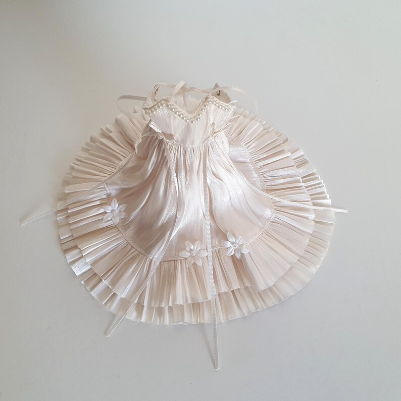 K&ouml;nnte beinhalten: Ein wei&szlig;es Satin-Kleid mit einem R&uuml;schenrock und Perlenverzierungen. Das Kleid hat einen schlichten Oberteil und wird an den Schultern mit B&auml;ndern gebunden.