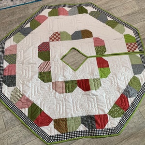 May include: A quilted Christmas tree skirt with a white center and a patchwork design in red, green, and gray. The skirt has a black and white checkered border with a green trim. The tree skirt is octagonal in shape.