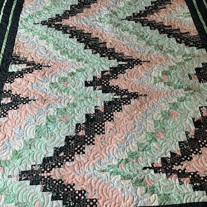 May include: A handmade quilt with a black border and a zigzag pattern. The quilt features sections of black polka dots, floral prints, and solid colors in shades of pink and green. The quilt is machine quilted with a wavy pattern.