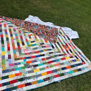 Könnte beinhalten: Handgefertigte Patchwork-Decke mit einem geometrischen Muster aus bunten Stoffstreifen. Die Rückseite der Decke zeigt ein Paisley-Muster. Die Decke liegt auf grünem Gras. Die Decke misst ca. 200 cm x 200 cm.
