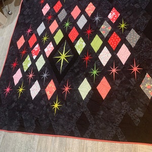 May include: A handmade quilt with a black background and a pattern of colorful diamonds and starbursts. The diamonds are in shades of red, pink, green, and gray, with floral and geometric patterns. The quilt is trimmed with a red border.
