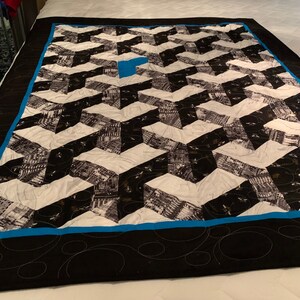 Könnte beinhalten: Eine geometrische Steppdecke mit einem schwarz-weiß-blauen Design. Die Steppdecke weist ein sich wiederholendes Muster aus schwarzen und weißen Formen auf, mit einem blauen Quadrat in der Mitte. Der Rand ist schwarz mit einem blauen Akzent und dekorativen Nähten.