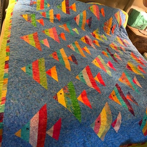 May include: A colorful quilt featuring a school of fish swimming across a blue background. The fish are made of various patterned fabrics in shades of red, orange, yellow, green, and blue. The quilt has a green border.