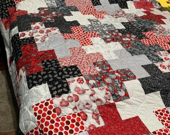 Puede incluir: Un edredón hecho a mano con un diseño de patchwork con telas rojas, negras, blancas y grises. El edredón tiene un patrón de cruz repetido, con varios patrones que incluyen lunares, flores y adornos navideños. El edredón está sobre una cama con un cabecero de mimbre.