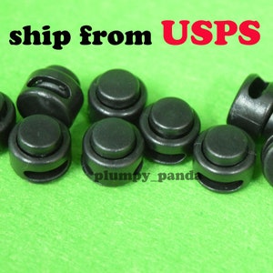 May include: A close-up of seven black plastic toggle stoppers. The stoppers have a round shape with a small hole in the center. The stoppers are arranged in a random pattern on a green surface.