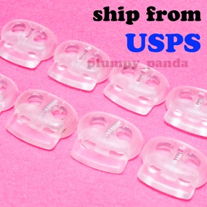 May include: Eight clear plastic skull-shaped toggle buckles. The buckles have a metal screw in the center and two holes on either side. The buckles are arranged in a row on a pink surface.