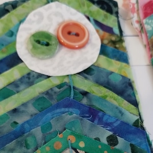 May include: Close-up of a handmade fabric art piece with a chevron pattern in shades of blue and green. A white fabric circle with a green and orange button is at the top. The piece is likely part of a larger quilt or textile project.