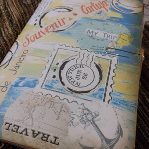 Pode incluir: Um diário de viagem com um design vintage. A capa apresenta selos, mapas e as palavras "Souvenir", "Travel" e "My Trip". A paleta de cores inclui azul, amarelo e bege, com um clip de papel prateado.