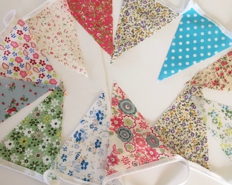 May include: A colorful bunting banner with triangular fabric flags. The flags feature various floral patterns in shades of pink, blue, green, and purple, with a light blue polka dot flag. The flags are strung together with white cording.