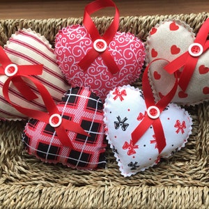 May include: Five red and white fabric heart-shaped ornaments with red ribbon bows and buttons. The hearts are arranged in a wicker basket.