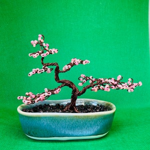 May include: A wire bonsai tree with pink beads representing blossoms, displayed in a blue and white ceramic pot. The tree is set against a green background.